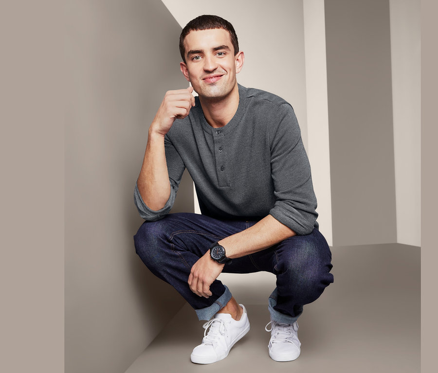 Muž v sivom žakárovom tričku henley pózuje v džínsoch a bielych topánkach.