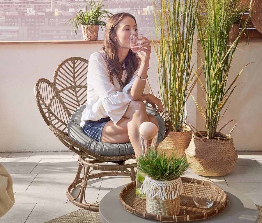 Žena sedí na otočnom pletenom kresle »Flower« a pije vodu. V pozadí je záhradná lavica a lounge kreslo »Boho«.