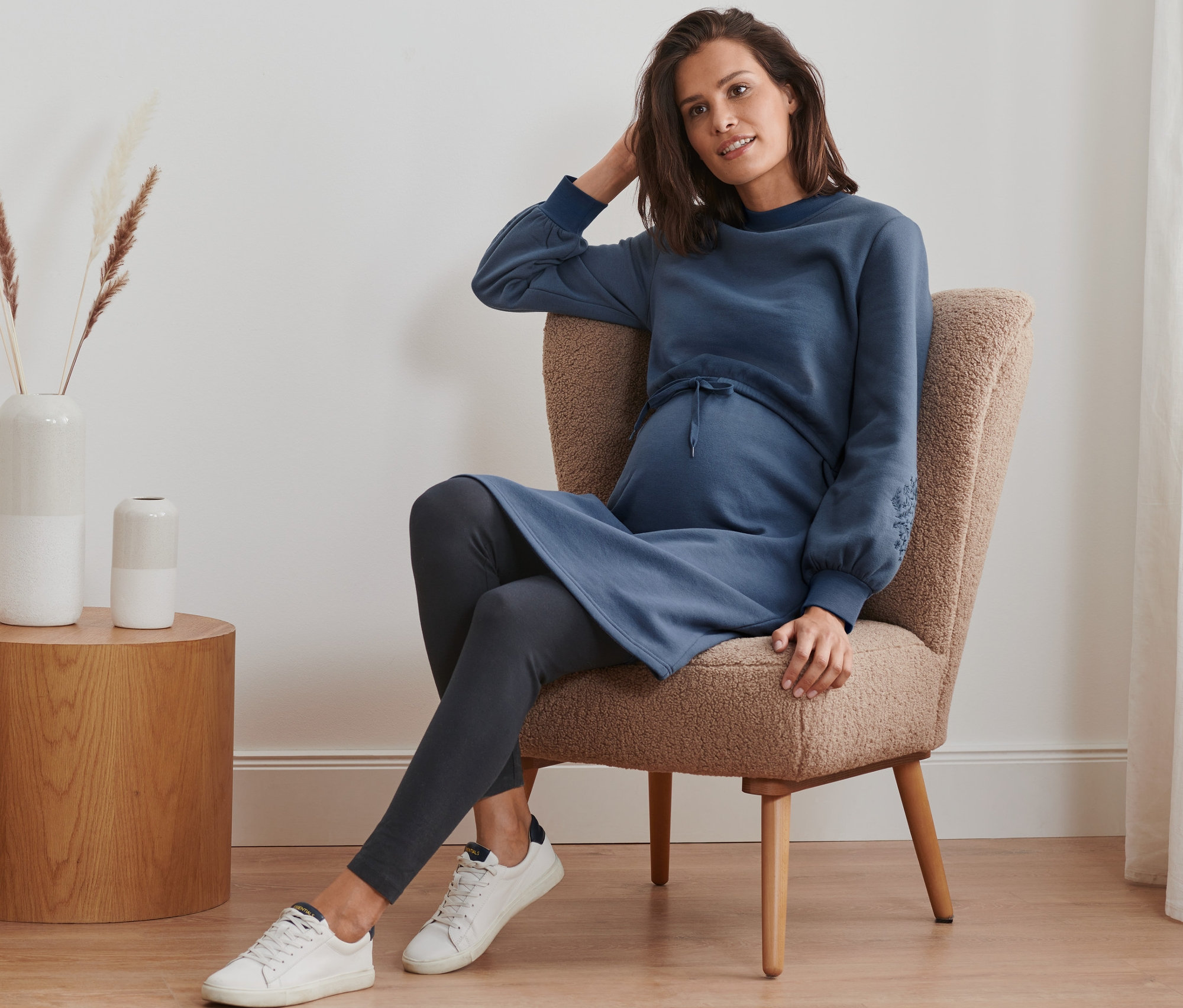 Tehotná žena sedí v kresle a má na sebe tehotenské a dojčiace teplákové šaty, legíny a tenisky.