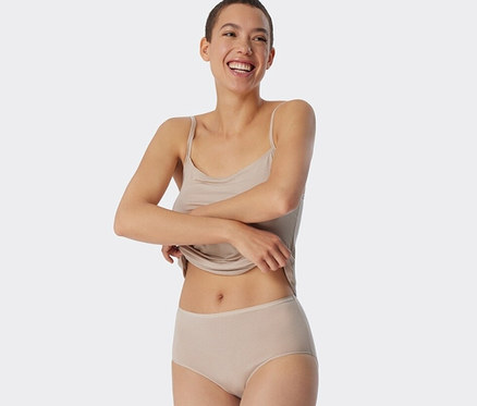 Žena sa usmieva a drží lem tielka. Má na sebe pieskové nohavičky SCHIESSER Midislip s prúžkami.