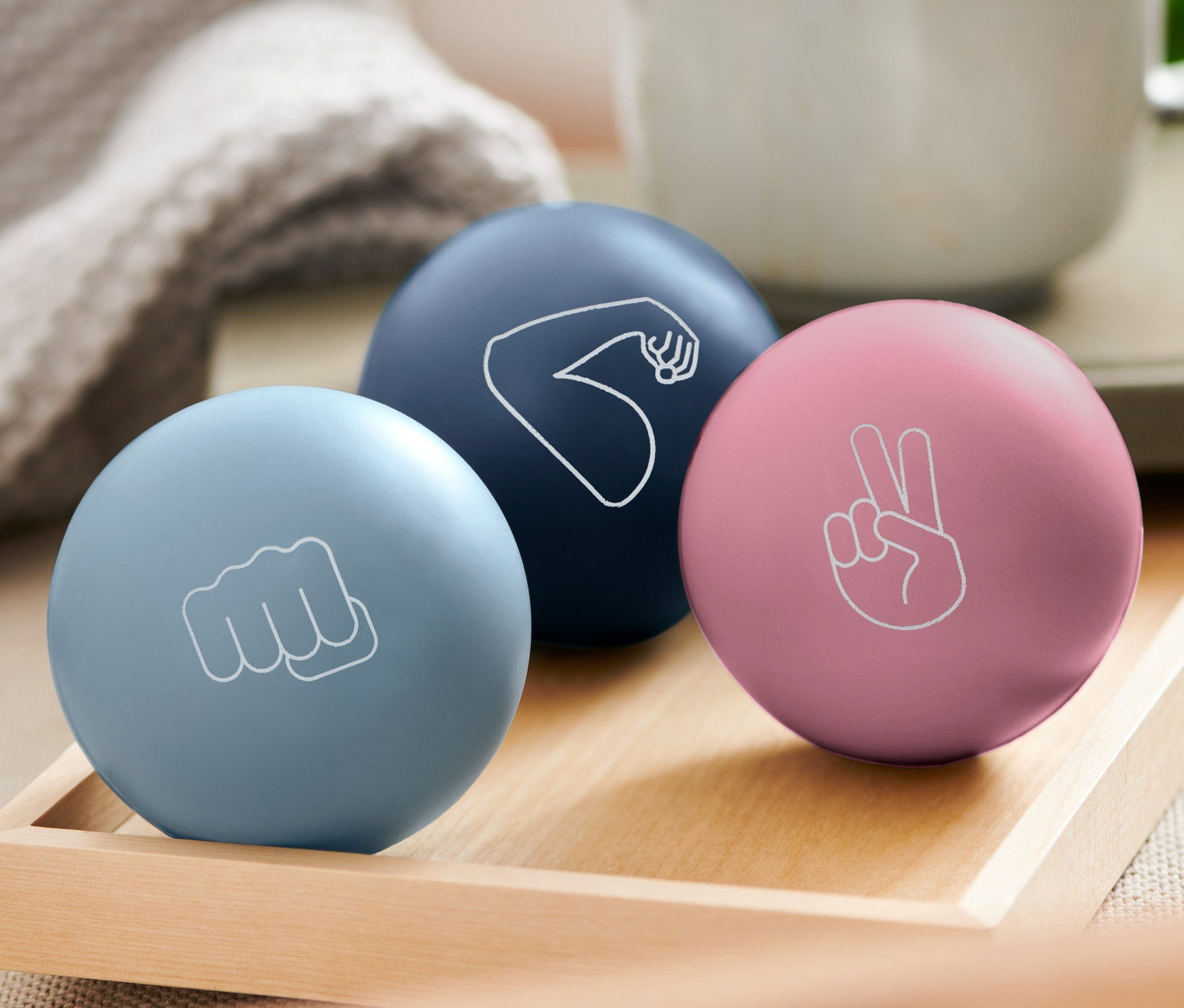 Na drevenom podnose ležia tri antistresové loptičky: svetlomodrá s päsťou, tmavomodrá s ramenom a ružová so znakom mieru. V pozadí je hrnček.