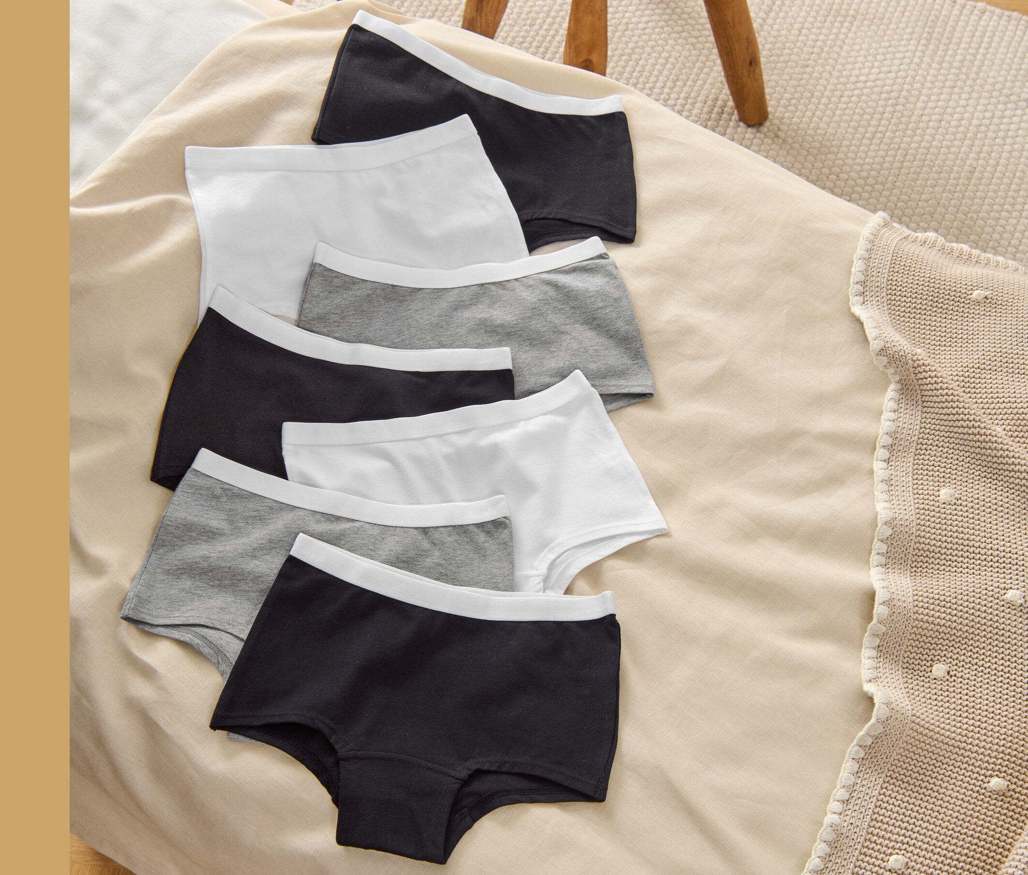 Sedem detských nohavičiek (Detské nohavičky, 7 ks, sivé, biele/čierne) leží na béžovej deke vedľa drevenej stoličky.