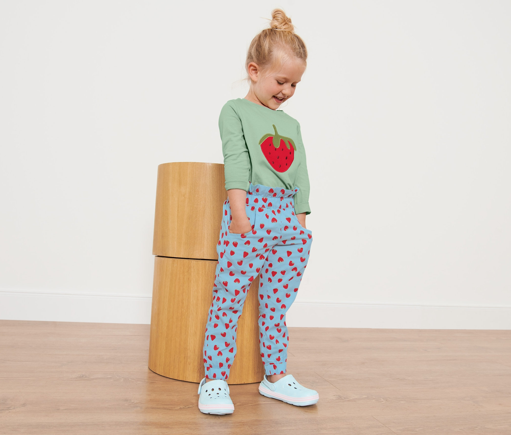 Dievča stojí a má na sebe detské nohavice v štýle paper bag s celoplošnou potlačou s motívom jahody.