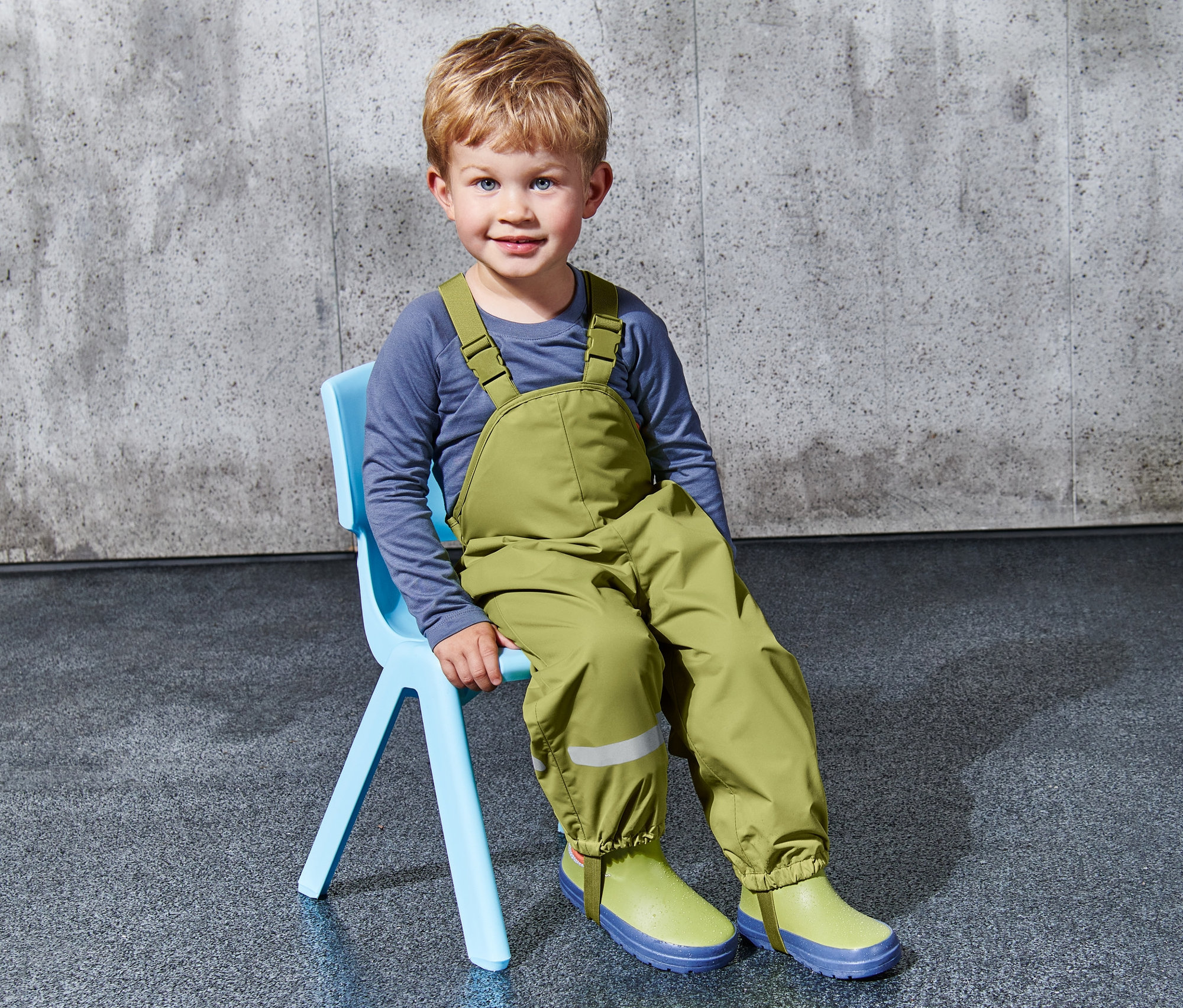 Chlapec sedí na modrej stoličke a má na sebe zelené detské nohavice do dažďa.