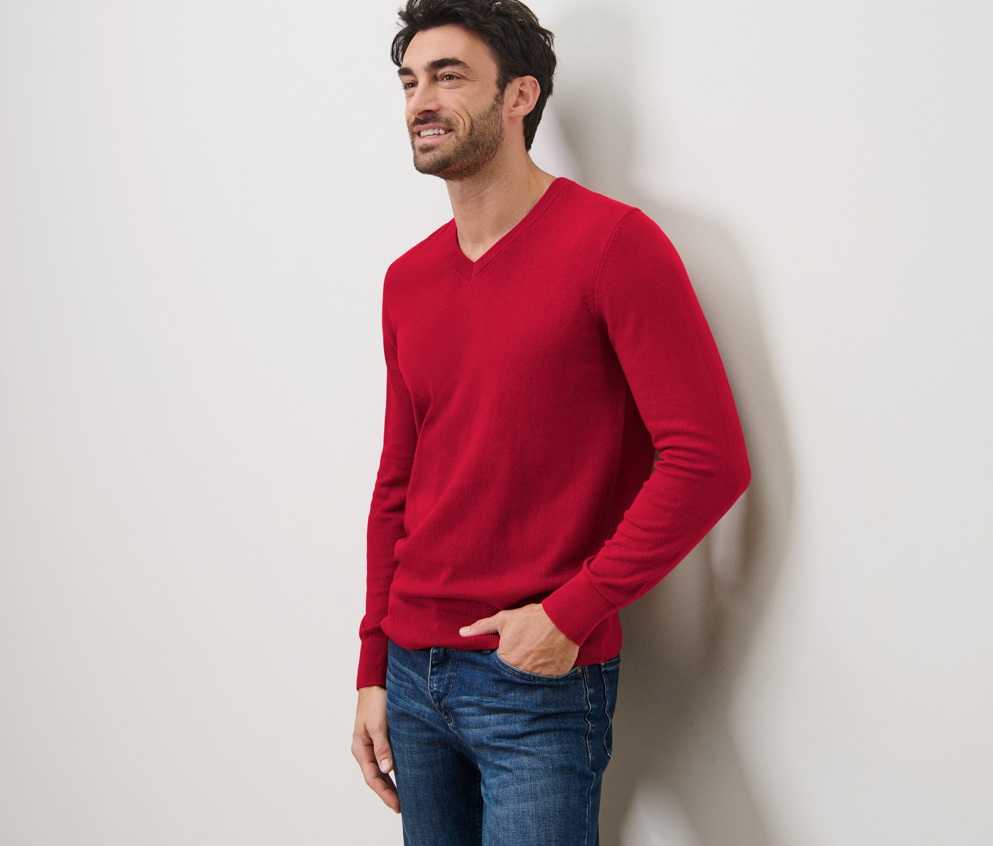 Muž sa usmieva a opiera sa o stenu. Má na sebe červený pulóver s výstrihom do V a džínsy.