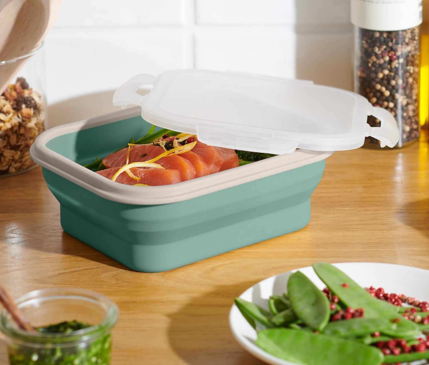 Tyrkysová skladacia dóza na potraviny s pootvoreným vekom, vnútri je losos. Na stole sú aj ďalšie potraviny.