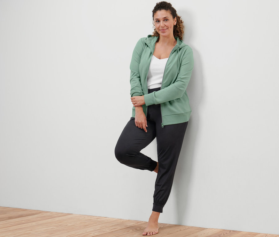 Žena opretá o stenu má na sebe šalviovozelenú mikinu a nohavice na jogu.
