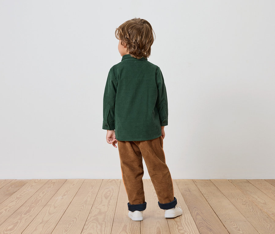 Chlapec v zelenej košeli a hnedých nohaviciach stojí chrbtom k divákovi na drevenej podlahe.