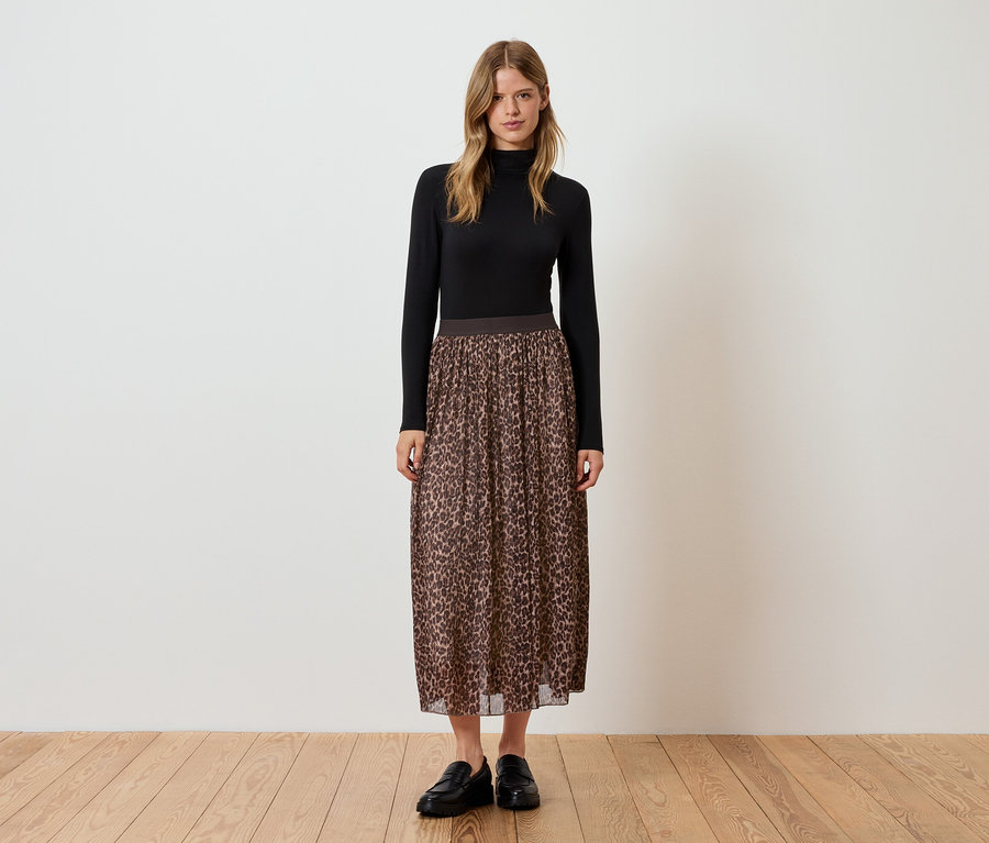 Žena stojí a má na sebe čierny rolák, hnedú skladanú sukňu so zvieracím vzorom a čierne topánky.