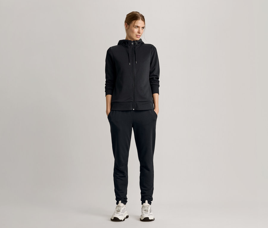 Žena stojí s rukami vo vreckách, oblečená v čiernej teplákovej súprave s mikinou s kapucňou a teniskami.