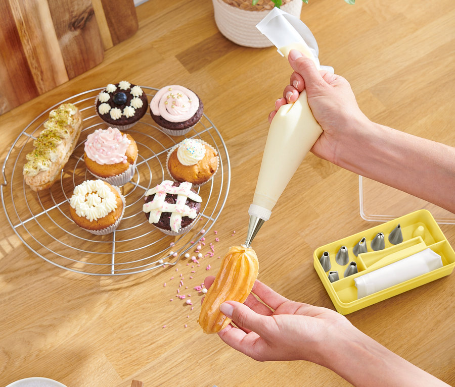 Osoba zdobí pečivo pomocou cukrárskeho vrecka, cupcakes a žltá krabička s tryskami ležia na drevenom stole.