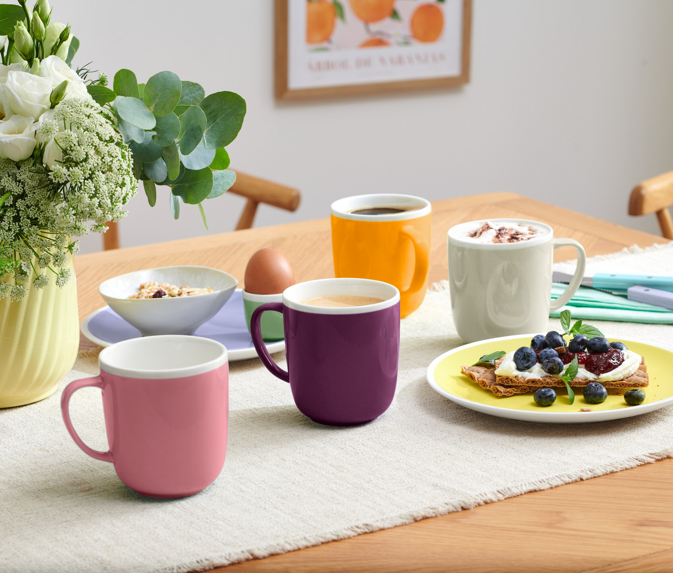 Na stole sú štyri hrnčeky rôznych farieb, misa s müsli, vajce v držiaku a tanier s toastom s čučoriedkami.