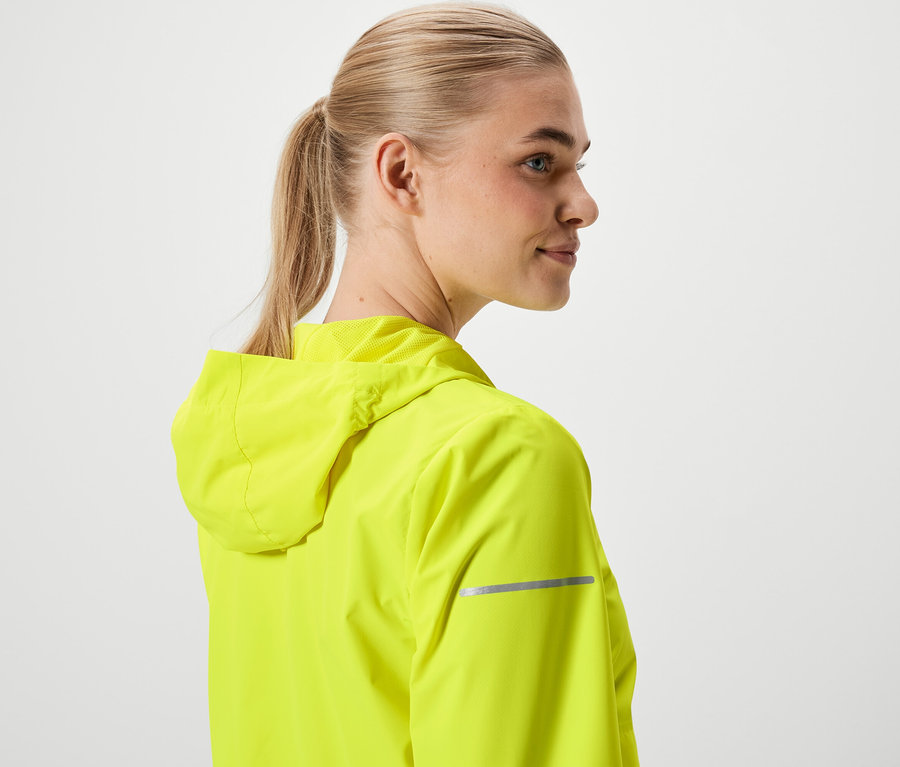 Žena s blond vlasmi stiahnutými do copu, oblečená v neónovo žltej bunde s kapucňou.