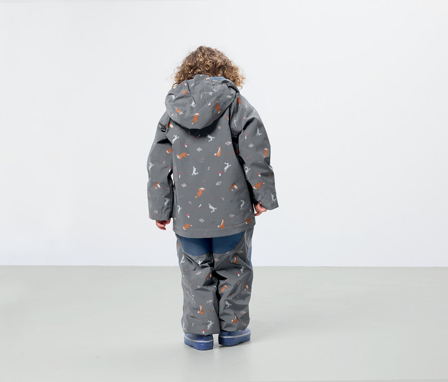 Chlapec s kučeravými vlasmi má na sebe sivú bundu a nohavice s potlačou líšok a vtákov, stojí chrbtom k fotoaparátu.