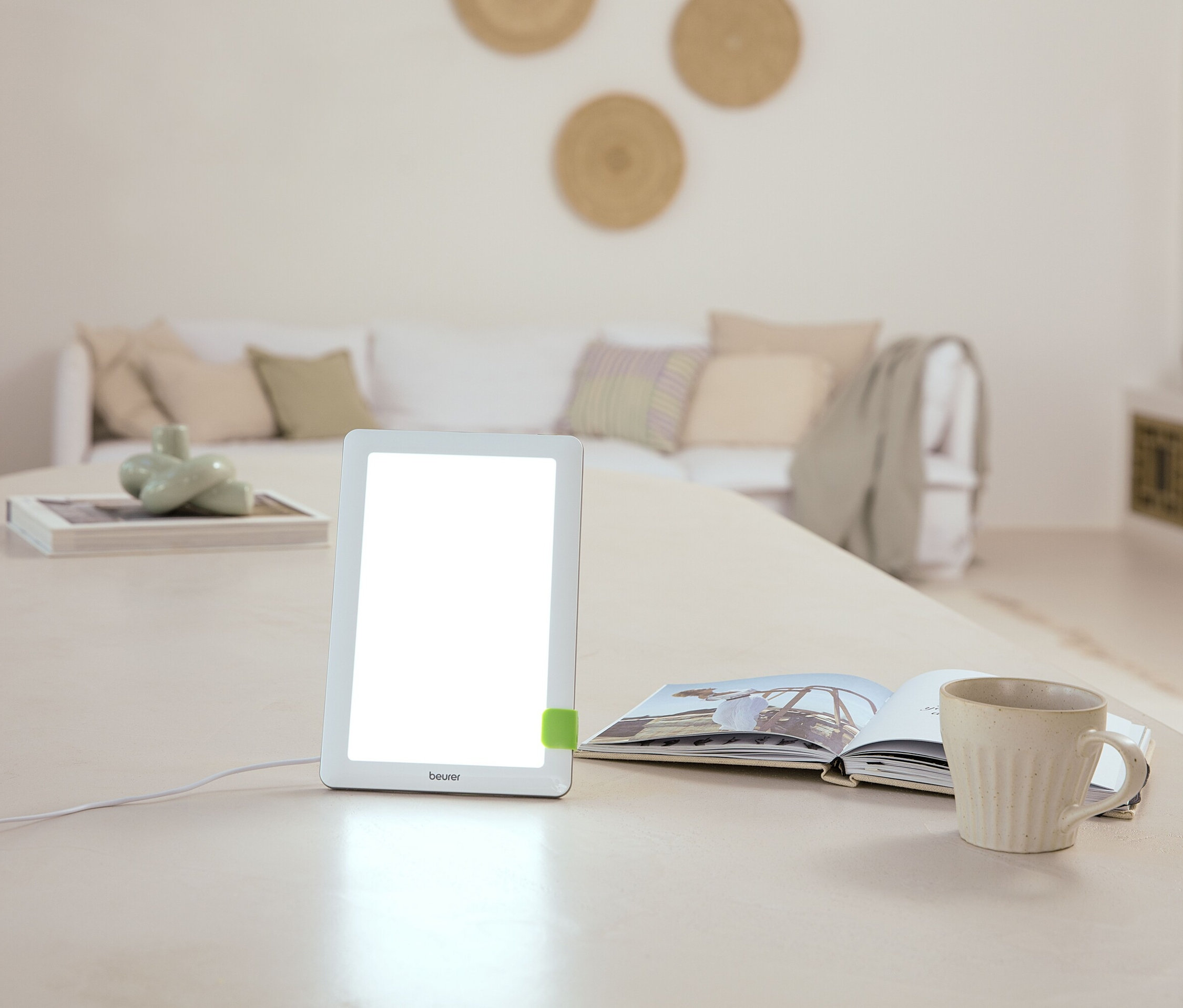 Na stole stojí lampa, otvorená kniha a hrnček. V pozadí je pohovka.