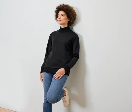 Žena s afro účesom sa opiera o bielu stenu. Má na sebe čierny rolák, modré džínsy a béžové tenisky.
