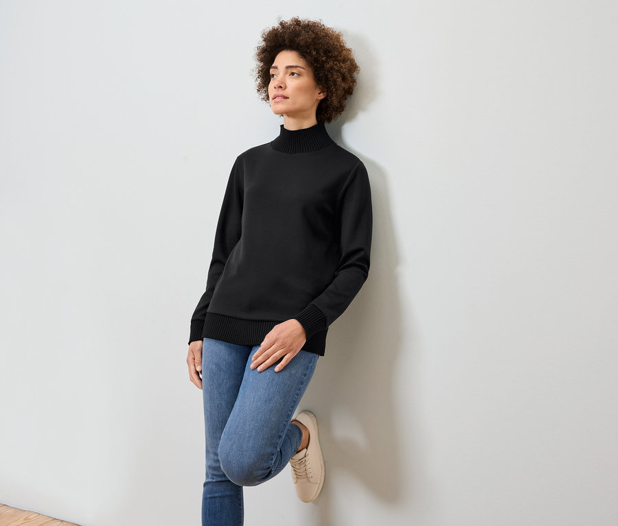 Žena s afro účesom sa opiera o bielu stenu. Má na sebe čierny rolák, modré džínsy a béžové tenisky.