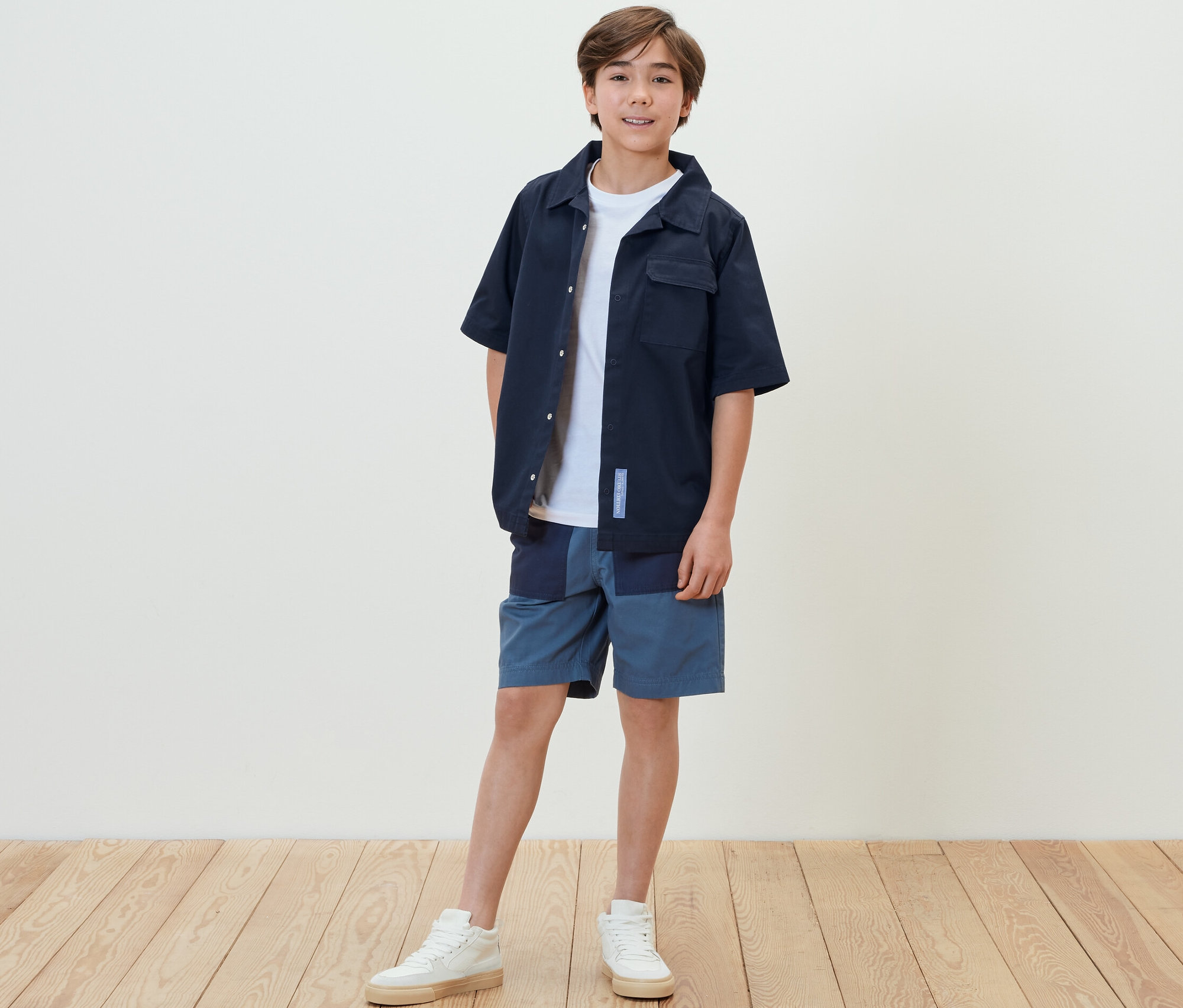 Chlapec stojí a má na sebe detskú bundu v košeľovom štýle, šortky a tenisky.