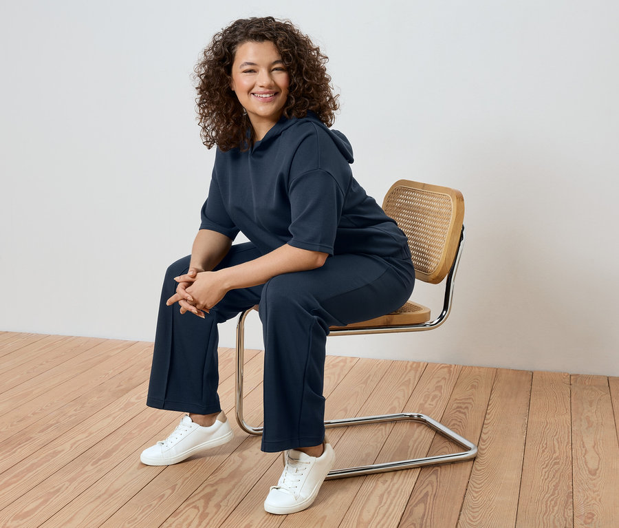 Žena s kučeravými vlasmi sedí na stoličke, oblečená v tmavomodrej teplákovej súprave a bielych teniskách.
