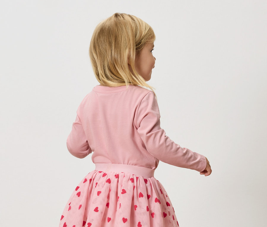Malé blond dievča má na sebe ružové tričko s dlhým rukávom a ružovú tylovú sukňu s červenými srdiečkami.