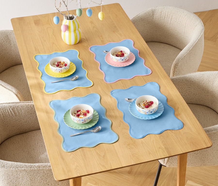 Jedálenský stôl je prestretý pre štyri osoby s miskami cereálií a farebnými podložkami, obklopený štyrmi stoličkami.