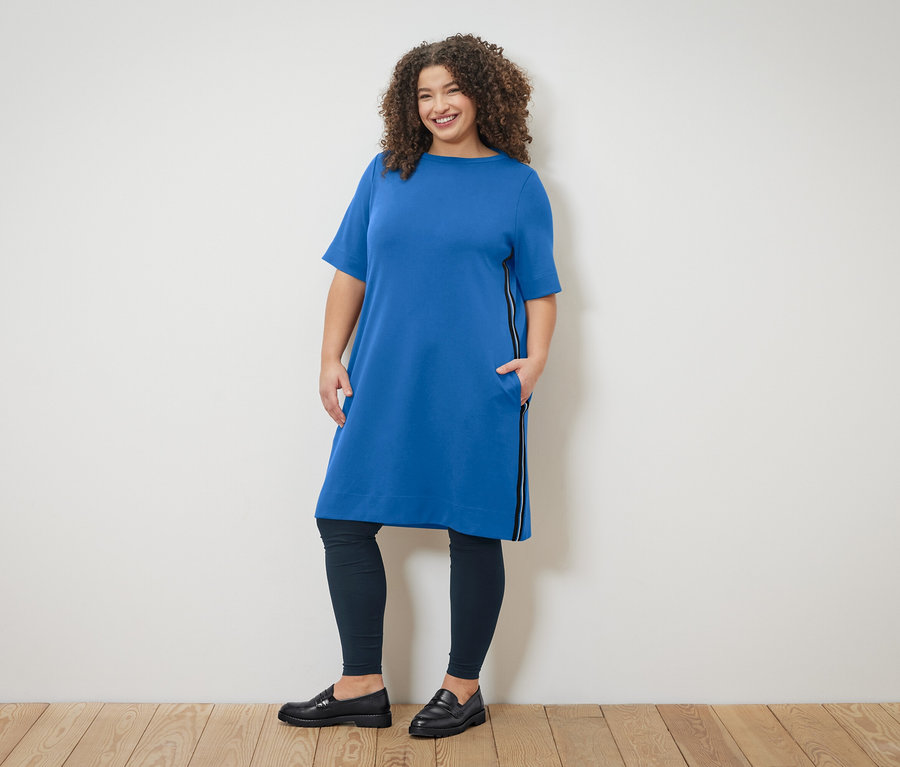 Žena s kučeravými vlasmi v modrých šatách so zipsom a legínach pózuje pred stenou.