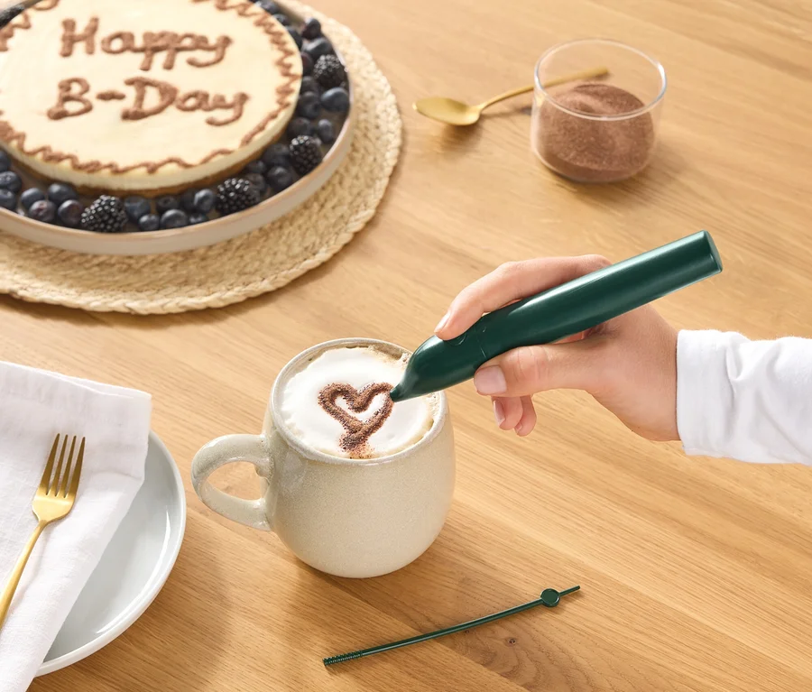 Ruka kreslí zeleným dekoračným perom kakaové srdce na penu kávy v hrnčeku. Na drevenom stole je tiež vidieť tortu s nápisom „Happy B-Day“ a bobuľami, pohár kakaového prášku, zlatú lyžičku a tanier s obrúskom a vidličkou.