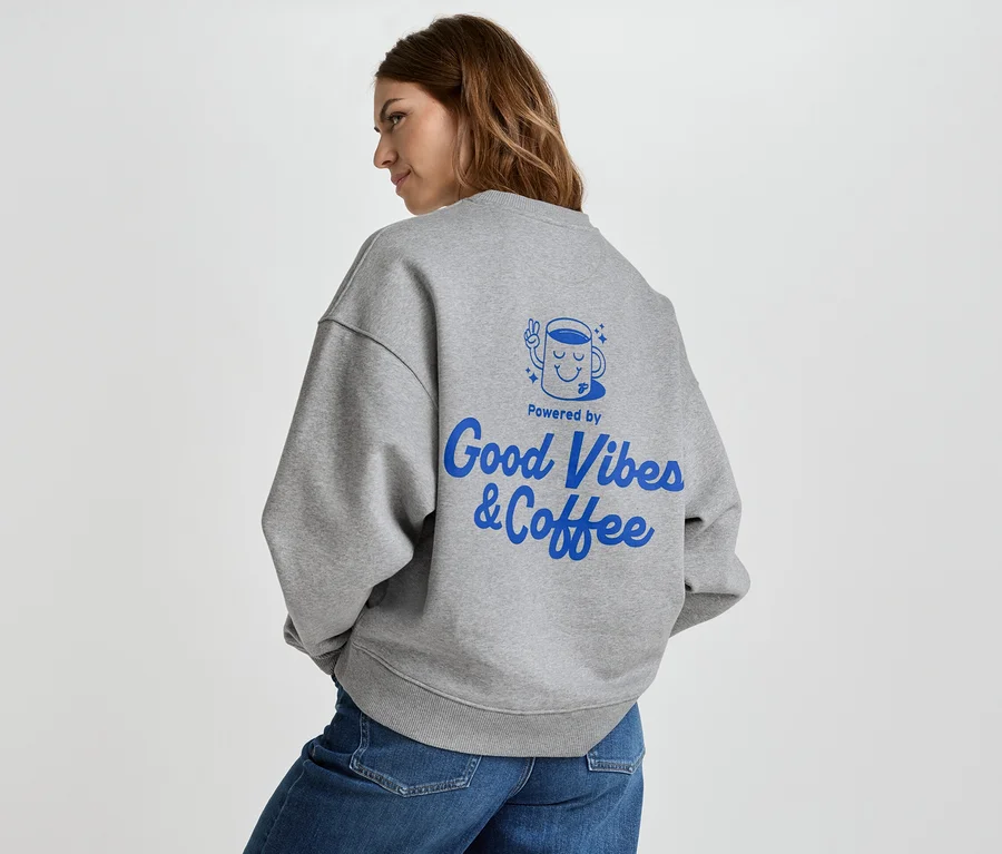 Žena má na sebe sivú mikinu s modrým nápisom "Good Vibes & Coffee" na chrbte a džínsy.