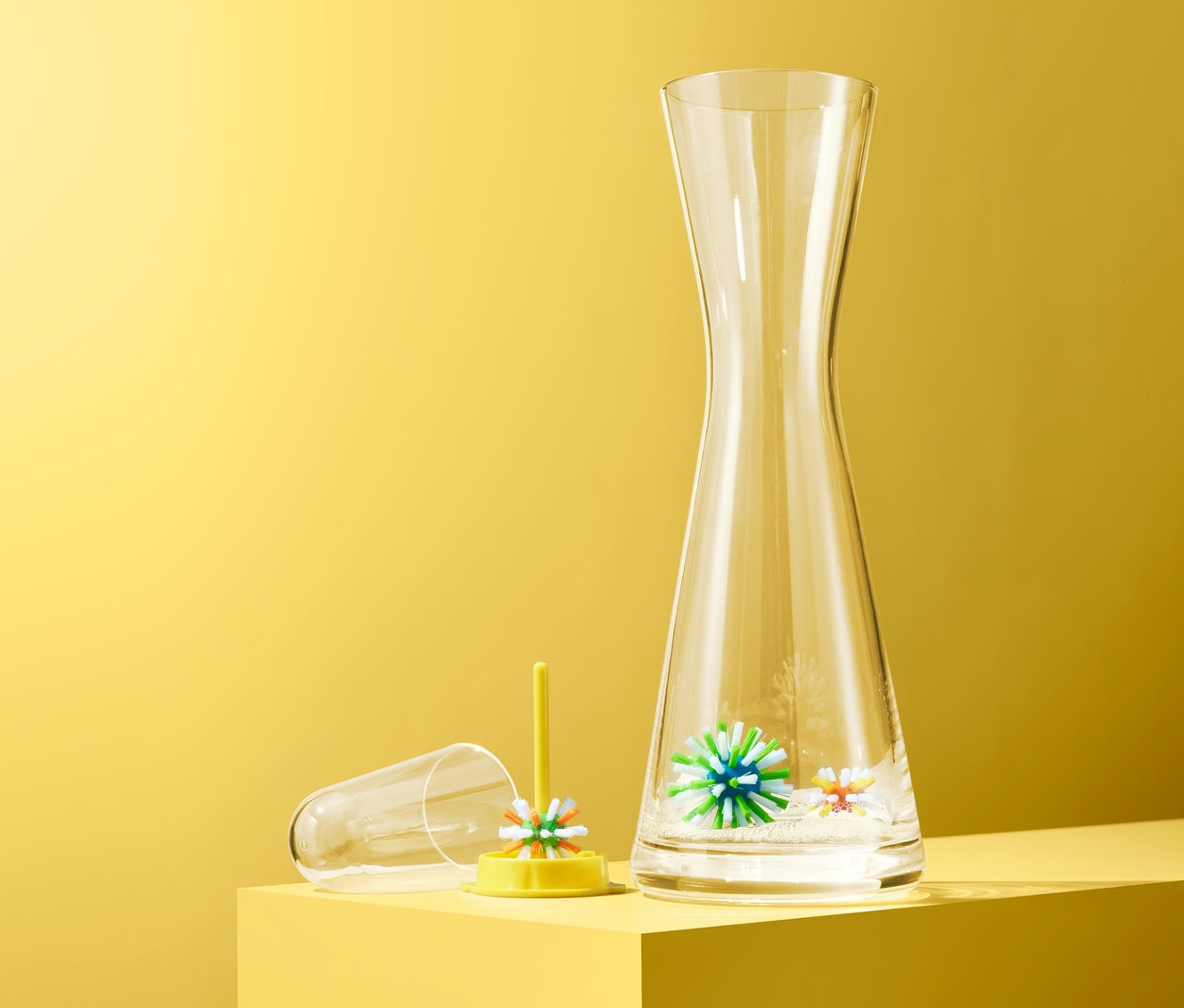 Číra karafa stojí vedľa žltého držiaka s loptičkami na čistenie fliaš, 3 ks, na žltom pozadí.