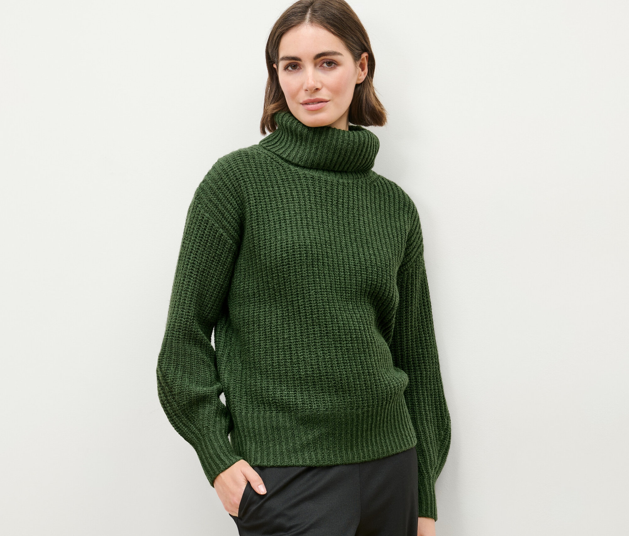 Žena pózuje na svetlom pozadí v pulóvri z hrubej pleteniny s rolákovým golierom.