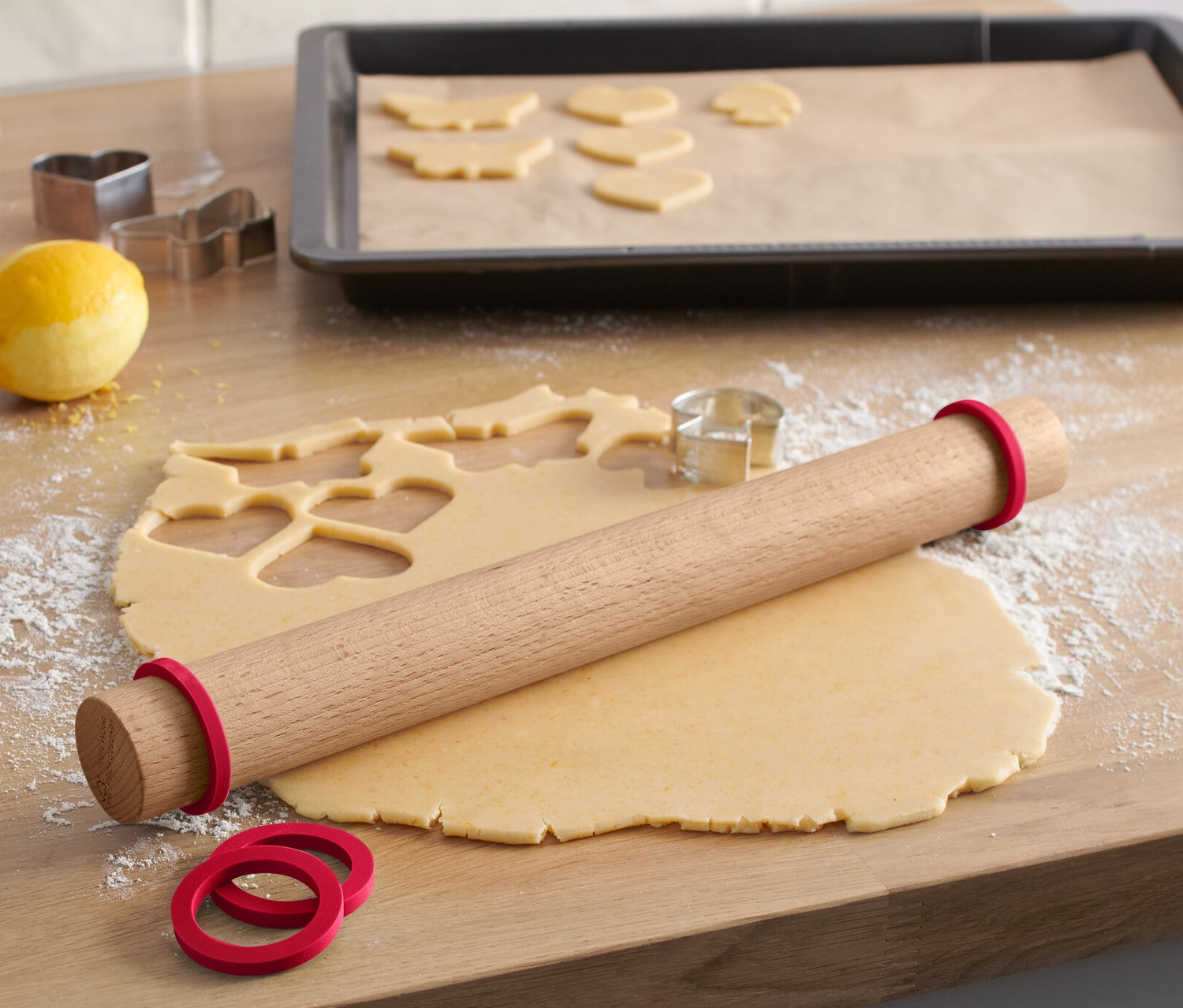 Na drevenej doske leží drevený valček na cesto s červenými gumičkami, vedľa je rozvaľkané cesto s vykrojenými tvarmi a formičky na vykrajovanie.