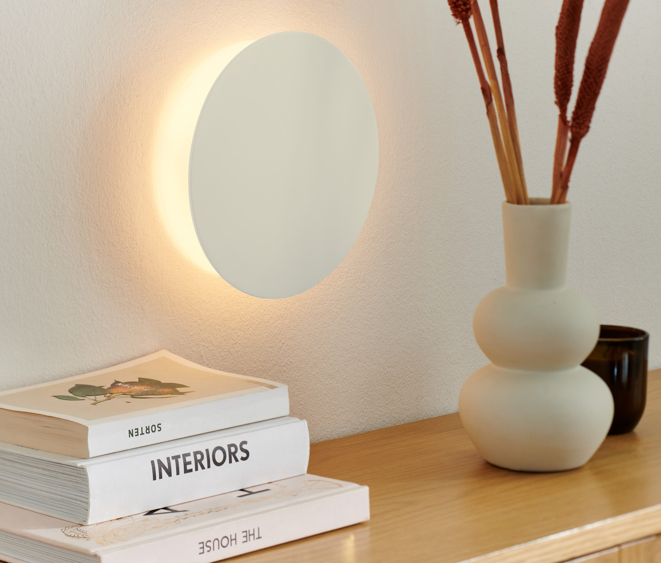 Na stene je umiestnené nástenné LED svietidlo na batérie. Na drevenej komode leží váza s okrasnými vetvičkami a knihy.