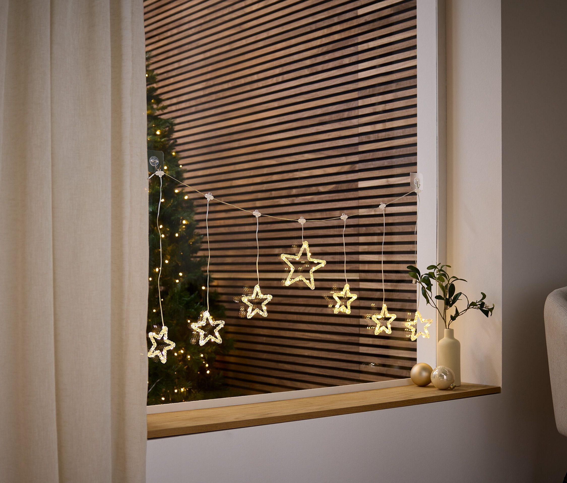 Reťaz svetiel v tvare hviezdy visí v okne, za ktorým je vidieť vianočný stromček.