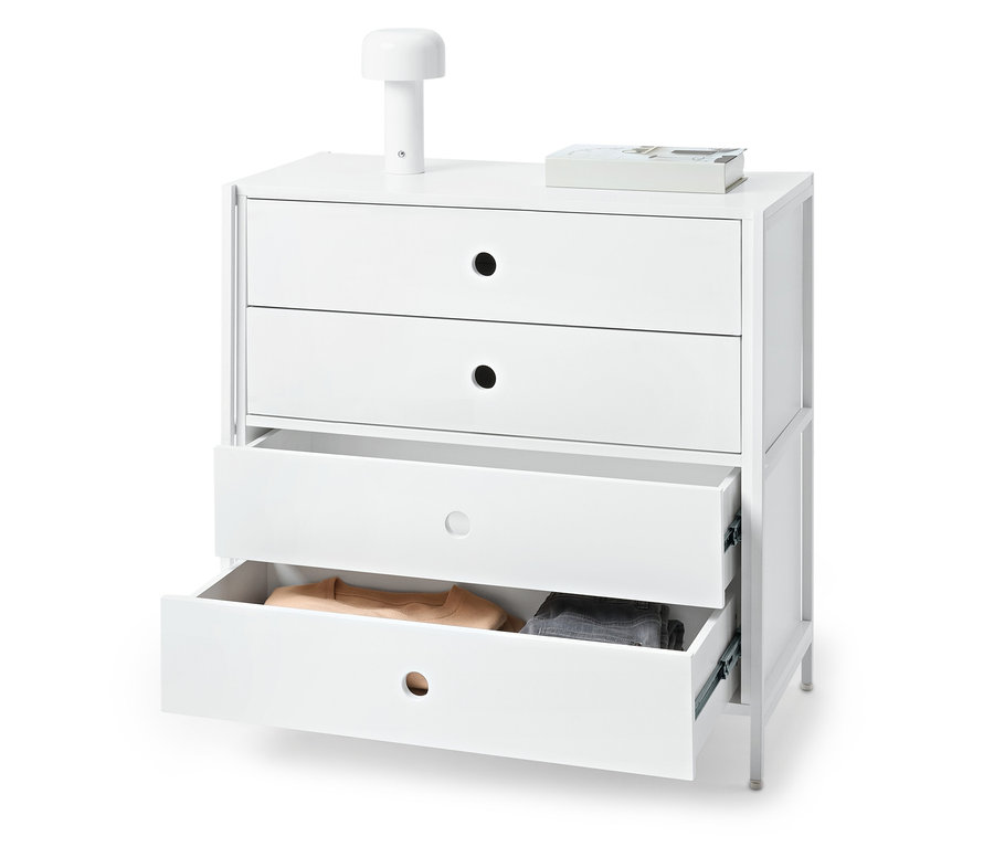 Biela komoda s tromi zásuvkami, pričom dve sú otvorené a obsahujú oblečenie. Na komode stojí lampa a kniha.