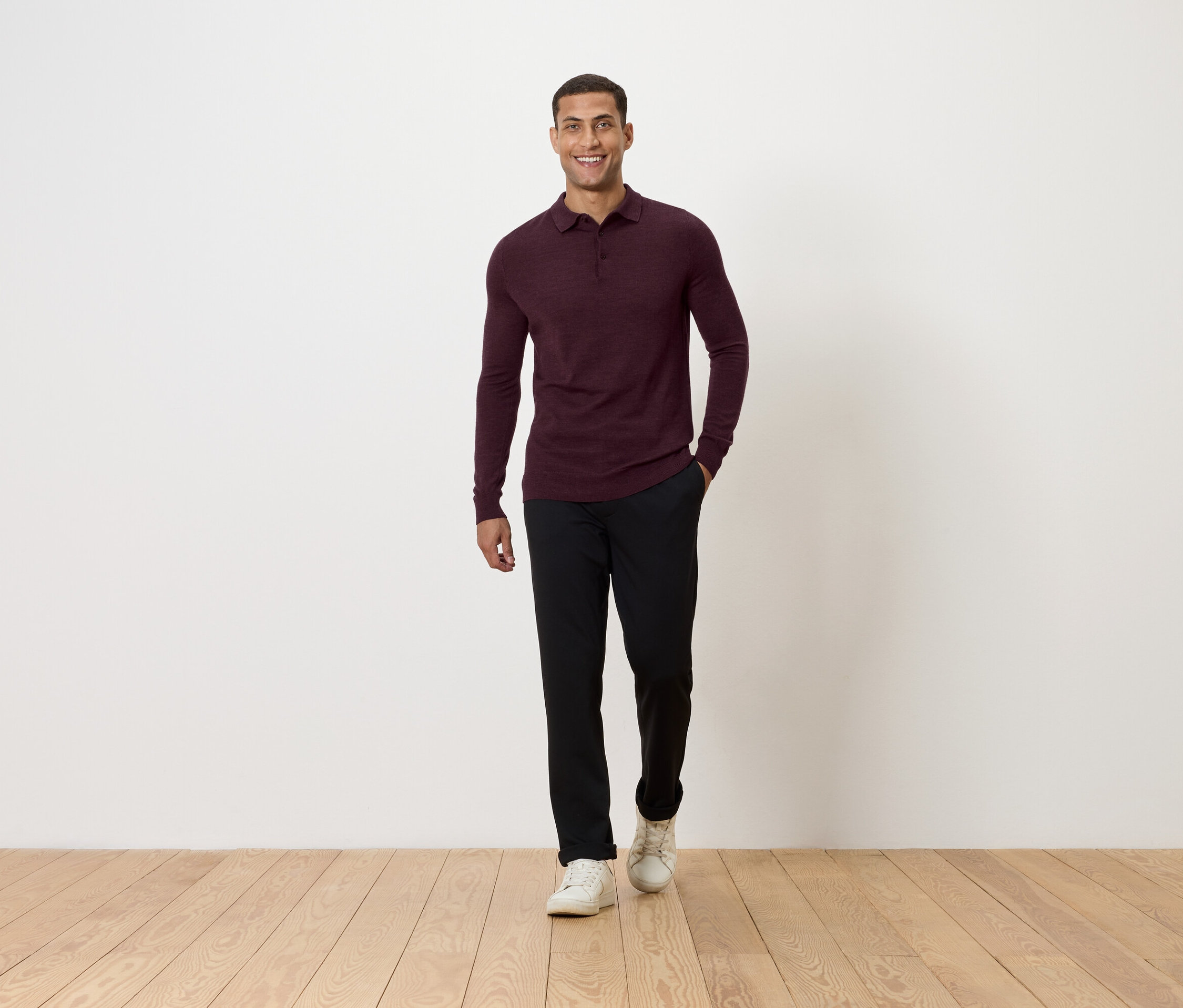 Muž ide po drevenej podlahe. Má na sebe bordové polo tričko s dlhým rukávom, čierne nohavice a biele tenisky.