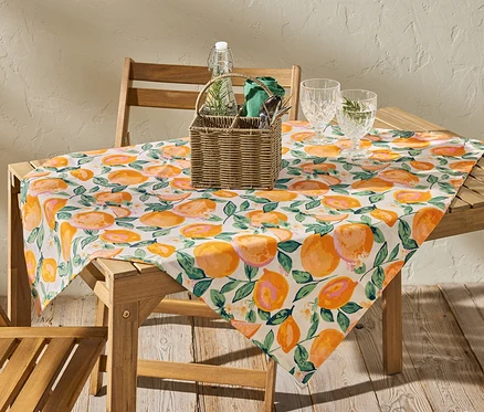 Na drevenom stole leží umývateľný obrus s potlačou oranžových citrusov, cca 90 x 90 cm.