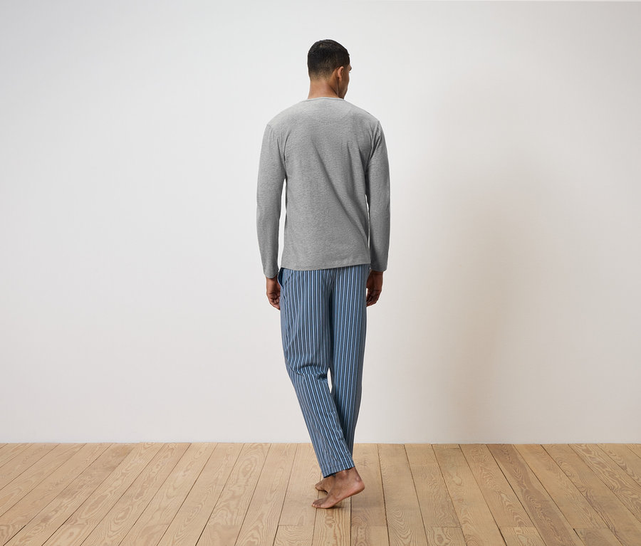 Muž chrbtom k fotoaparátu stojí na drevenej podlahe. Má na sebe sivé tričko s dlhým rukávom a modré pruhované pyžamové nohavice.