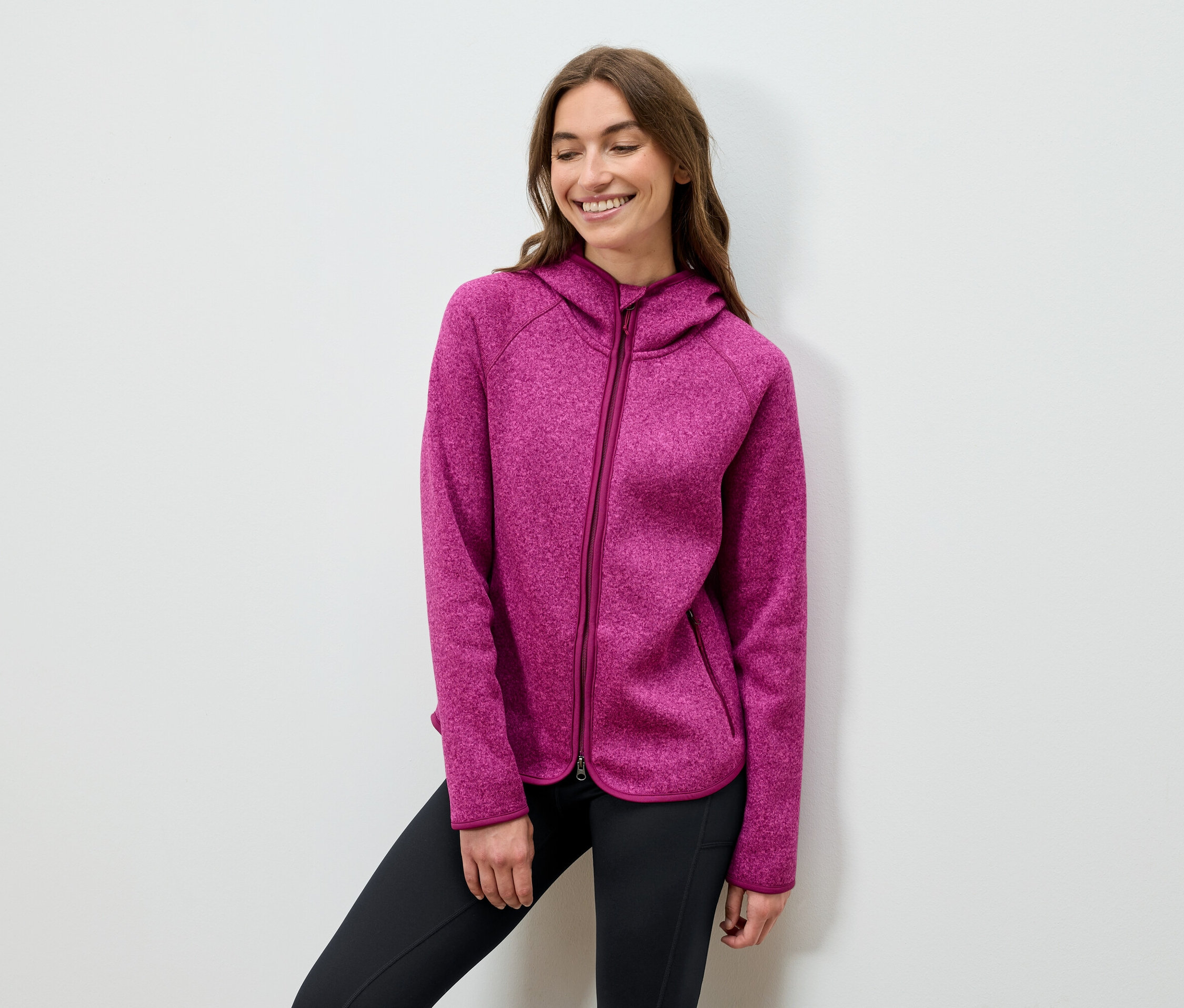 Usmievajúca sa žena pózuje v ružovej mikine s kapucňou a čiernych nohaviciach.