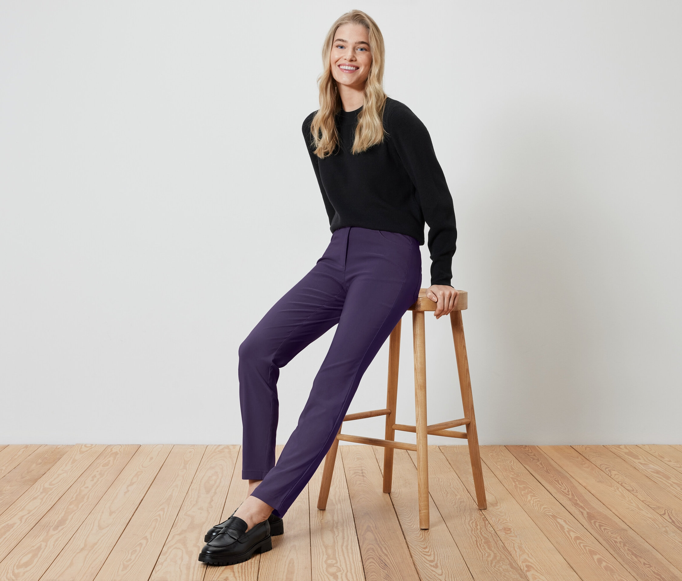 Blondínka sedí na drevenej stoličke, má na sebe čierne tričko a fialové nohavice.