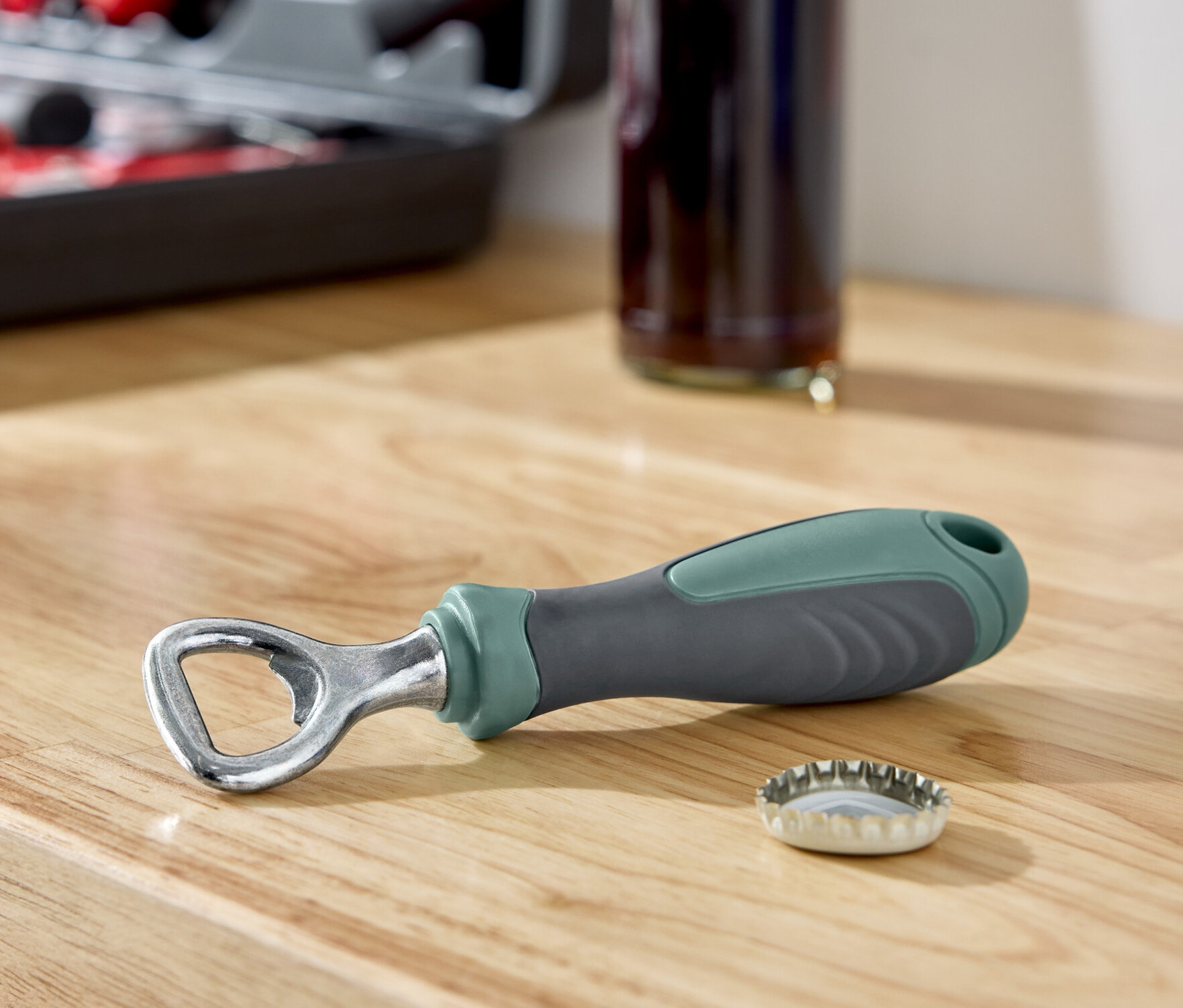 Na drevenej doske leží otvárač na fľaše vedľa viečka, v pozadí je fľaša a sada náradia.