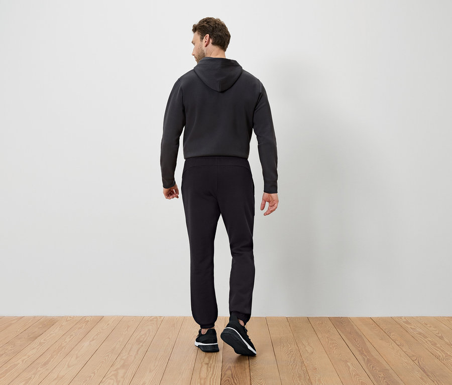 Muž chrbtom k fotoaparátu má na sebe tmavú mikinu s kapucňou a čierne tepláky.