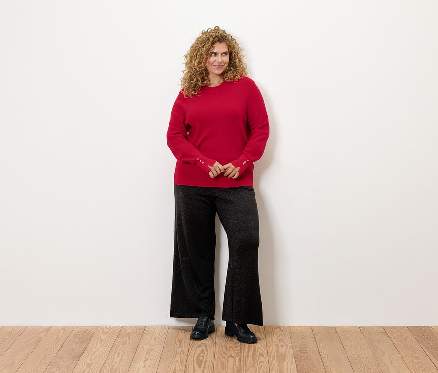 Žena s kučeravými vlasmi stojí a má na sebe červený sveter a čierne nohavice.