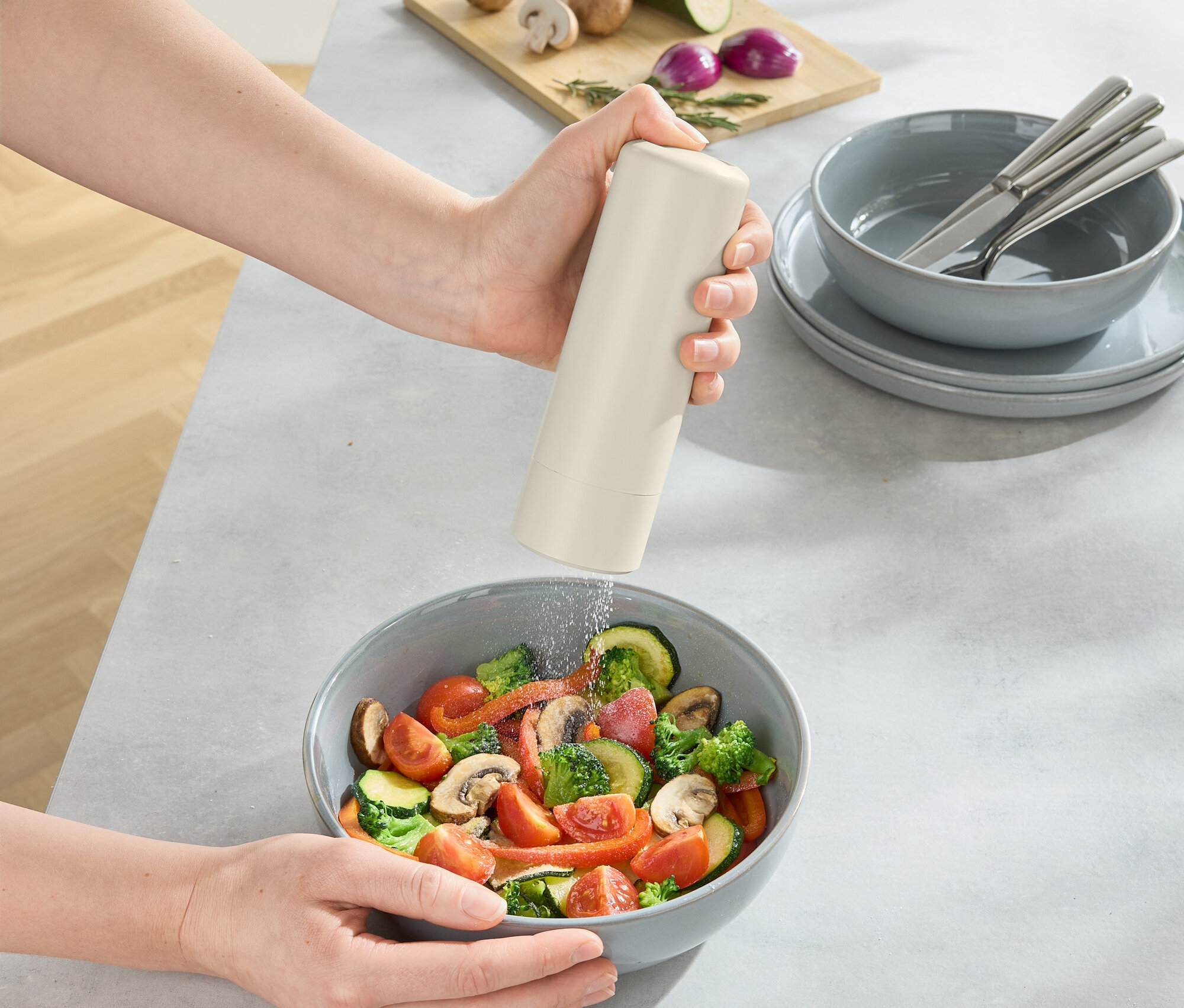 Osoba korení misku šalátu, na stole je doska s hubami, cibuľou a cuketou.