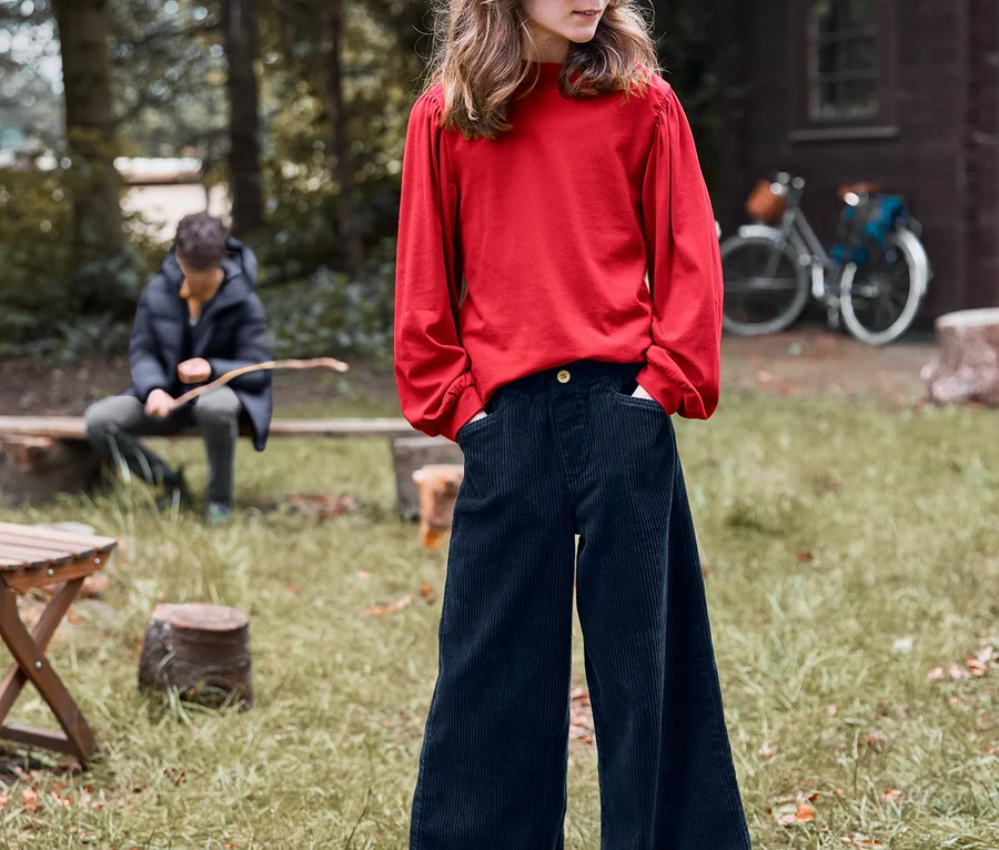 Dievča stojí vonku a má ruky vo vreckách menčestrových nohavíc culottes a na sebe tričko s dlhým rukávom.