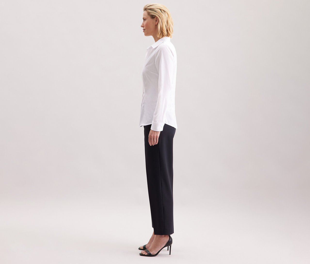 Profil ženy nosí bielu blúzku Seidensticker Slim Fit non iron, kombinovanú s tmavými nohavicami a čiernymi vysokými podpätkami.