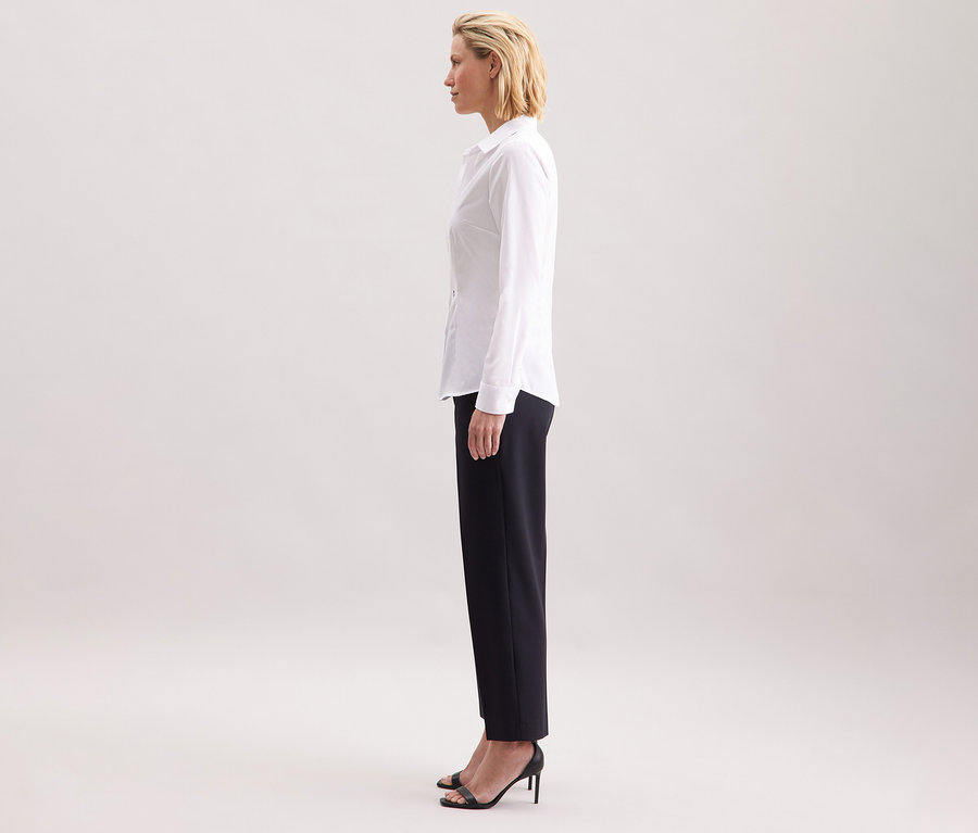 Profil ženy nosí bielu blúzku Seidensticker Slim Fit non iron, kombinovanú s tmavými nohavicami a čiernymi vysokými podpätkami.