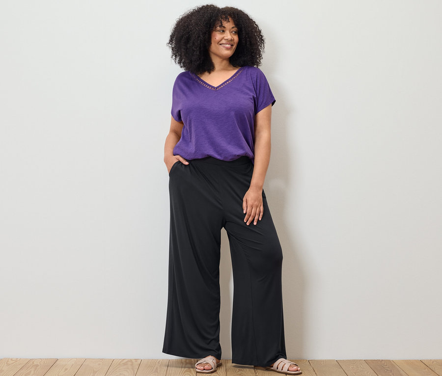 Žena pózuje v fialovom tričku zo zmesi modalu a čiernych nohaviciach v štýle palazzo.