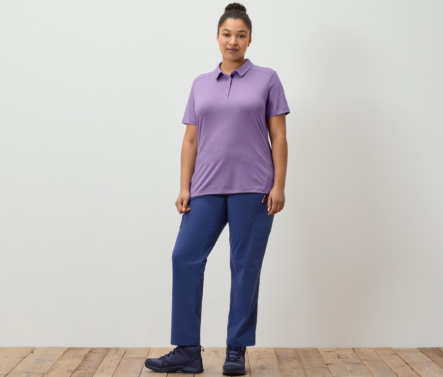 Žena pózuje na drevenej podlahe v tmavomodrých outdoorových nohaviciach a fialovej funkčnej polokošeli.