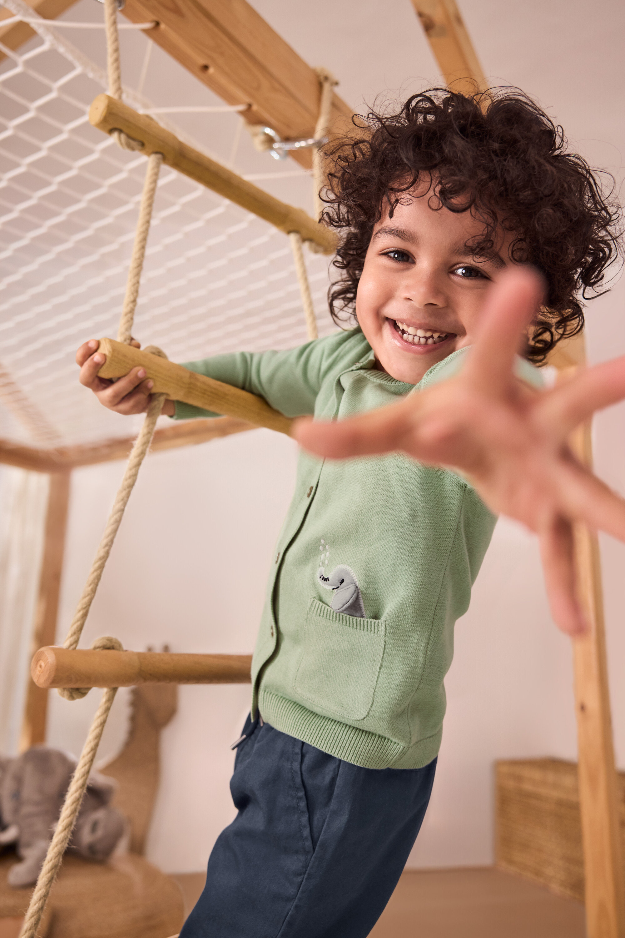 Usmievavý chlapec lezie po povrazovom rebríku. Má na sebe zelený detský pletený sveter s aplikáciou a modré detské naťahovacie nohavice – strih »Anton«.