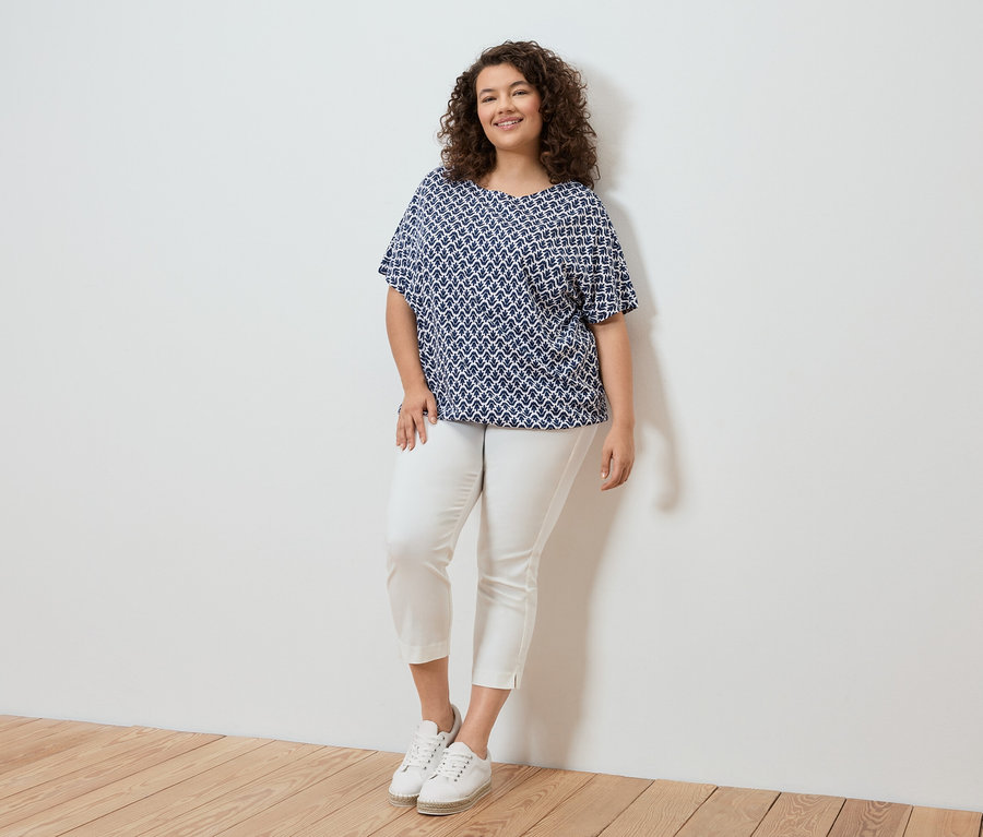 Žena s kučeravými vlasmi v modro-bielej blúzke so vzorom, bielych nohaviciach a bielych teniskách stojaca pred bielou stenou.